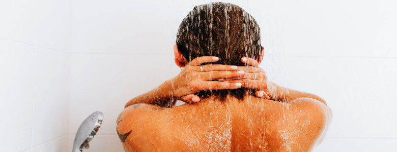 Oberer Rücken einer blonden Frau unter der Dusche mit Tattoo am linken Oberarm